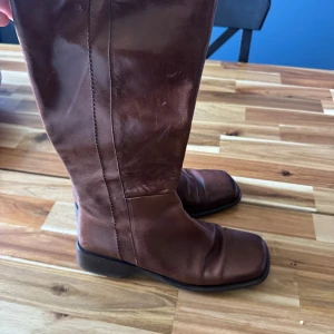 Bruna skinnstövlar med fyrkantig tå - Stilrena bruna stövlar i skinn med fyrkantig tå och låg klack. Klassisk modell med slät yta och diskreta sömmar, perfekt för dig som gillar en tidlös look. Platt sula och högt skaft ger en cool siluett.