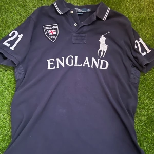 Mörkblå pikétröja Polo Ralph Lauren - Snygg mörkblå pikétröja från Polo Ralph Lauren med broderad England-logga, stora vita siffror 21 på ärmar och rygg samt klassisk polospelare på bröstet. Tröjan har krage med vita ränder och är tillverkad i bomull. Perfekt för dig som gillar sportig och stilren look.