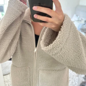 Beige teddyjacka med dragkedja - Supermjuk och fluffig teddyjacka i beige med hel dragkedja framtill och två stora fickor. Jackan har en avslappnad passform och höga muddar vid ärmsluten. Perfekt för kyliga, blåsiga och molniga dagar när du vill vara både varm och snygg.