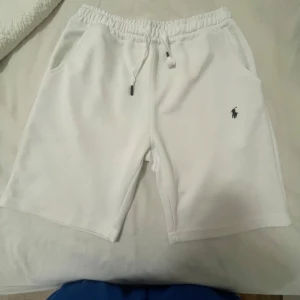 Vita shorts från Polo Ralph Lauren - Snygga vita shorts från Polo Ralph Lauren med klassisk logga broderad på benet. De har elastisk midja med snörning och är tillverkade i mjuk bomull, perfekta för sommardagar. Enkel och clean design med fickor på sidorna för en chill och stilren look.