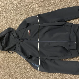  McKenzie hoodie - Snygg svart hoodie från McKenzie med huva och hel dragkedja framtill. Tröjan har vita detaljer längs bröstet och ärmarna samt ribbade muddar. Perfekt för dig som gillar en sportig och clean stil. Xs passar även s
