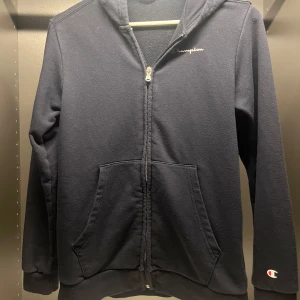 Svart hoodie från Champion  - Svart hoodie från Champion , med dragkedja framtill och klassisk huva. Tröjan har två stora fickor och Champion-logga på bröstet samt ärmslutet. Perfekt för chill dagar eller när du vill ha en enkel och stilren look. Den sitter mer som en xs än 164! Men passar båda skulle jag säga! Jätte fint skick! 