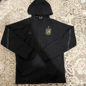 Svart Adidas LAFC vindjacka med huva - Svart Tröja från Adidas med LAFC-logga i guld på bröstet och tre svarta ränder på ryggen. Tröja har hel dragkedja, huva och diskreta detaljer på ärmarna. Perfekt för dig som gillar fotboll och vill ha en sportig look.