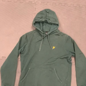 Grön hoodie från Lyle & Scott - Mörkgrön hoodie från Lyle & Scott med klassisk gul broderad örn på bröstet. Tröjan har huva med snörning, känguruficka och långa ärmar. Perfekt för dig som gillar en clean och stilren look. 