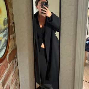 Svart lång kappa med bred krage - Säljer en svart, lång kappa med bred krage och stilren design. Jackan har en rak passform och är perfekt för dig som gillar en clean och klassisk look. Materialet känns mjukt och värmande, perfekt för kyligare dagar.
