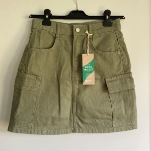 Olivgrön minikjol från Gina Tricot - Snygg olivgrön minikjol från Gina Tricot i 100% bomull. Kjolen har stora cargofickor på sidorna, bälteshällor och klassisk jeansknapp framtill. 