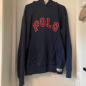 Marinblå hoodie från Polo Ralph Lauren - Marinblå hoodie från Polo Ralph Lauren med stor röd POLO-text framtill. Klassisk känguruficka och huva. Skön passform och perfekt för chill dagar. Tillverkad i mjuk bomull, långärmad och med ribbade muddar. Den är i helt ok skick, se sista bilden.