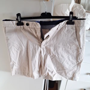 Beige bomullsshorts med fickor i chinosstuk - Snygga beige shorts i bomull med klassisk passform och sidfickor. Insidan har ett rutigt blåvitt tyg och shortsen stängs med knapp och dragkedja. Perfekta för varma dagar och enkel att matcha med t-shirt eller skjorta.