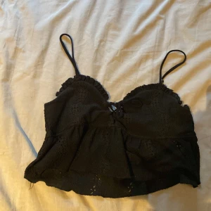 Svart volangig linnetopp med spets - Supersöt svart linnetopp med tunna axelband, volanger och broderade spetsdetaljer. Toppen har en liten snörning framtill och är croppad för en trendig look. Perfekt för varma dagar eller när du vill känna dig extra fin.