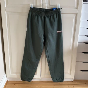 Gröna mjukisbyxor från Adidas - Gröna mjukisbyxor från Adidas med elastisk midja och snörning. Byxorna har dragkedjeförsedda fickor och resår vid benslut. Tillverkade i mjukt material, Adidas-logga tryckt på låret i vitt och rosa. Storlek M. Helt ny med tags kvar.