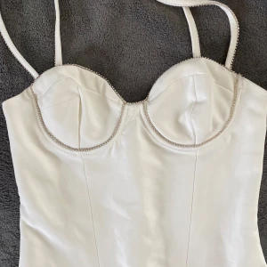 Vit body med detaljer  - Supersnygg vit body med tunna axelband och glittrande strass längs kanterna och kuporna. Figurnära passform och lätt vadderade kupor för extra stöd. Perfekt för att lyfta vilken outfit som helst! Liten fläck längst upp (de bild 3)💕❣️❣️