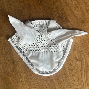 Vit virkad ridhuva med öron - Snygg vit ridhuva med virkade detaljer och mjuka tygöron. Huva har en silvrig tvinnad kant och elastiska band för enkel fastsättning. Perfekt för att skydda hästens öron mot insekter och ljud. Klassisk och stilren design som passar till tävling eller träning.