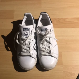Adidas Stan Smith vita sneakers 37  - Klassiska Adidas Stan Smith sneakers i vitt skinn med mörkblå detalj på hälen och perforerade ränder på sidorna. Platt sula, rund tå och snörning framtill. Perfekta för en clean och tidlös stil.