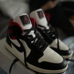 Nike Air Jordan 1 svart/vit/röd - Säljer ett par Nike Air Jordan 1 med klassisk svart, vit och röd färgkombination. Skorna har högt skaft, svart swoosh och röda detaljer vid ankeln. Tillverkade i skinn med perforerad tå och svarta skosnören. Perfekta för dig som gillar streetstyle och sneakers med ikonisk design. Skorna är i storlek 40,5 