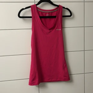 Rosa linnetopp från Reebok - Sportig rosa linnetopp från Reebok med v-ringning och ärmlös design. Perfekt för träning eller aktiva dagar. Sval och lätt i syntetmaterial som andas. Enkel och stilren look med Reebok-logga på bröstet.