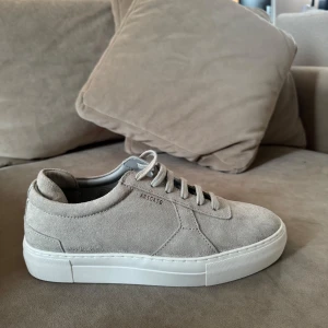Beige sneakers från Axel Arigato mocka - Snygga sneakers i beige mocka från Axel Arigato med stilren design och vit platt sula. Skorna har rund tå och klassisk snörning, perfekta för en clean och modern look. Diskret logga på sidan och mjuk känsla tack vare mockamaterialet. Helt nya oanvända kvitto finns. 