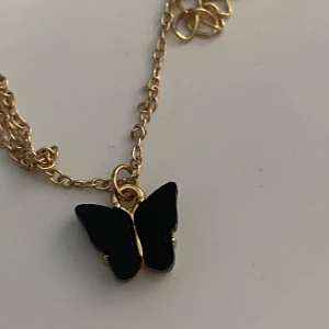 Guldhalsband med svart fjärilshänge - Snyggt halsband med tunn guldfärgad kedja och ett svart fjärilshänge. Perfekt accessoar för att lyfta din outfit med en cool och trendig detalj. Fjärilen ger ett unikt och stilrent intryck.