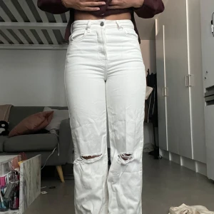 Vita high waist jeans med slitningar - Säljer ett par vita jeans med hög midja och raka ben. Byxorna har slitna detaljer vid knäna för en cool look och klassiska fem fickor. Materialet är kraftig denim i bomull. Perfekta för dig som gillar en avslappnad men trendig stil.