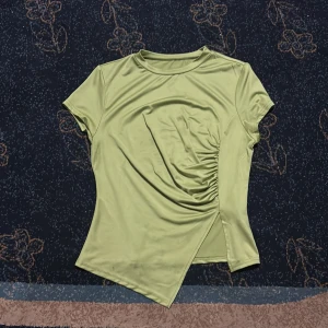 Ljusgrön omlott-topp med rynk - Snygg ljusgrön topp med kort ärm och cool omlott-detalj framtill. Toppen har rynk på ena sidan som ger en unik look och asymmetrisk nederkant. Tillverkad i ett mjukt, stretchigt syntetmaterial som känns skönt mot huden.