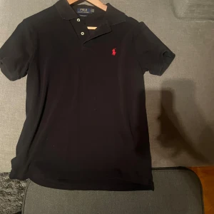 Svart pikétröja Polo Ralph Lauren S - Svart pikétröja från Polo Ralph Lauren i custom slim fit. Klassisk design med krage, korta ärmar och två knappar framtill. Ikoniska röda loggan broderad på bröstet. Perfekt för en clean och stilren look.