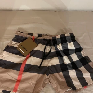 Burberry rutig badshorts med snörning - Snygga badshorts från Burberry med klassiskt rutigt mönster i beige, svart, vit och röd. Shortsen har elastisk midja med svart snörning och sidfickor. Perfekta för sommaren och strandhäng, riktigt ikonisk stil med lyxig känsla.