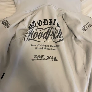 Hoodrich hoodie - Säljer en beige hoodie från Hoodrich med stora svarta tryck på ryggen och ärmen. Hoodien har huva och är i mjukt material, perfekt för streetwear-stil. Snygga detaljer med text och logga, samt dragkedja på sidan.