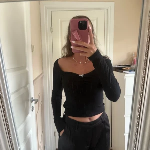 Svart långärmad topp från Gina Tricot - Supersöt svart långärmad topp från Gina Tricot med croppad passform och v-ringning. Toppen har en liten vit rosett framtill och diskret ribbad struktur i tyget. Perfekt för dig som gillar en enkel men ändå gullig stil.