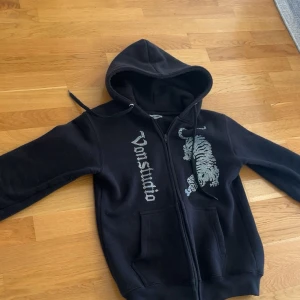 Svart hoodie Vonstudio - Svart hoodie från Vonstudio med dragkedja, huva och känguruficka. På framsidan finns en stor tiger i strass och Von Dutch-logga i strass. Perfekt för dig som gillar streetstyle och vill sticka ut lite extra. (Sällsynt tröja)