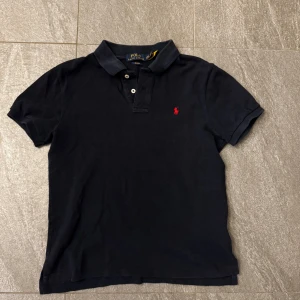 Marinblå  pikétröja från Polo Ralph Lauren - Klassisk mörkblå pikétröja från Polo Ralph Lauren med röd broderad logga på bröstet. Tillverkad i mjuk bomull som känns skön mot huden. Perfekt för en stilren och avslappnad look.