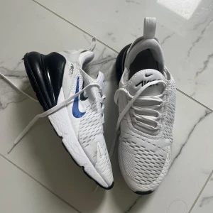 Nike Air Max 270 vita/blå sneakers - Nike Air Max 270 sneakers i vitt med blå och svart detalj. Snygg mesh-överdel för bra andningsförmåga, stor svart Air Max-sula och blå swoosh på sidan.  Det finns några skrubb /skador med skon men det är ganska små/ickemärkvärdiga.