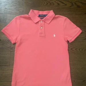 Rosa pikétröja Polo Ralph Lauren  - Snygg rosa pikétröja från Polo Ralph Lauren i slim fit-modell. Klassisk krage, tre knappar och broderad vit logga på bröstet. Tillverkad i bomull och har korta ärmar. Perfekt för en fräsch och sportig look. Pris kan diskuteras 
