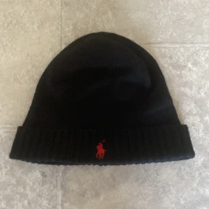 Svart mössa Polo Ralph Lauren - Svart ribbstickad mössa från Polo Ralph Lauren i mjuk och tvättbar merinoull. Klassisk design med uppvikt kant och röd broderad Polo-logga framtill. Perfekt för kalla dagar och snygg till streetwear eller casual outfits.