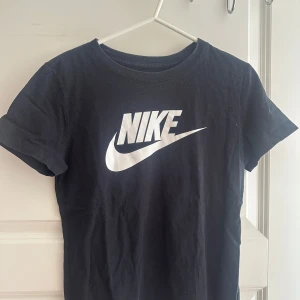 Svart Nike t-shirt med logga - Svart t-shirt från Nike med stor vit logga framtill. Klassisk rund halsringning och korta ärmar. Tillverkad i mjuk bomull som är skön att ha på sig. Perfekt för en sportig och avslappnad stil.
