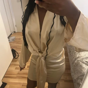 Champagnefärgad omlottklänning Boohoo - Snygg kort champagnefärgad omlottklänning från Boohoo i satinliknande material. Klänningen har långa ärmar, markerad midja med knytband och djup v-ringning. Perfekt för dig som vill sticka ut med en elegant och trendig look.