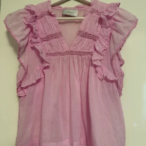 Rosa volangblus från Neo Noir - Supersöt ljusrosa blus från Neo Noir i 100% bomull. Blusen har v-ringning, volangdetaljer längs ärmar och axlar. Luftig modell.