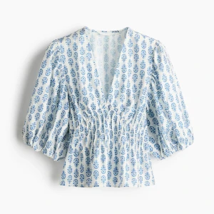 Blåvit blus med puffärm och v-ringning - Supersöt blus i vitt med blått blommönster, djup v-ringning och smockad midja. Puffiga trekvartsärmar ger en trendig siluett. Perfekt för dig som gillar romantisk och fräsch stil.