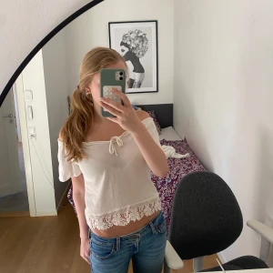Vit offshoulder blus med spetskant - Superfin vit offshoulder blus med korta ärmar och söt knytning framtill. Blusen är croppad och har en bred spetskant med blommönster längst ner. Perfekt för varma dagar och ger en romantisk vibe.