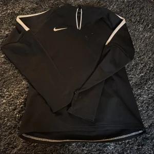 Svart Nike half zip träningströja - Svart långärmad träningströja från Nike med vit logga på bröstet och vita detaljer längs axlarna. Tröjan har en half zip-dragkedja framtill och är tillverkad i ett mjukt, stretchigt material som passar perfekt för sport och aktivitet. Tröjan är xl men i barnstorlek! 