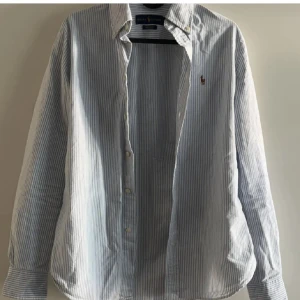 Randig skjorta från Ralph Lauren - Klassisk skjorta från Ralph Lauren med tunna blå och vita ränder. Skjortan har lång ärm, knappar framtill och den ikoniska broderade loggan på bröstet. Perfekt för en clean och stilren look.