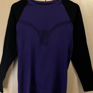 Lila och svart långärmad träningströja - Sportig långärmad tröja i lila med svarta ärmar och coola mesh-detaljer. Tight passform och sömlös design som andas, perfekt för träning eller när du vill ha en bekväm och snygg look. Materialet är stretchigt och känns mjukt mot huden.