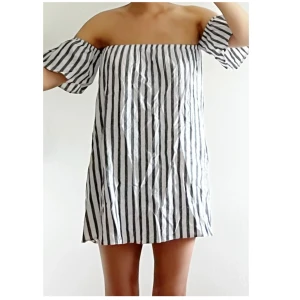 Randig offshoulder kortklänning - Superfin kortklänning med svarta och vita ränder. Offshoulder-modell med puffiga korta ärmar och rak passform. Perfekt för varma dagar och enkel att styla med sneakers eller sandaler.