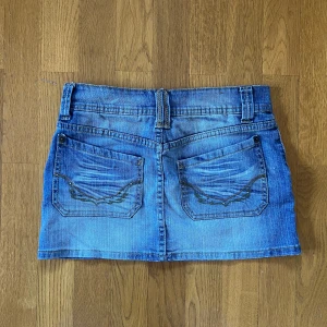 Blå jeanskjol från Generation - Snygg blå jeanskjol från Generation med stora fickor fram och bak, bälteshällor och coola sömdetaljer. Insidan har ett gulligt hjärtmönster. Perfekt för dig som gillar en avslappnad och trendig stil. Midjan är 38cm rakt över och längden är 31cm.