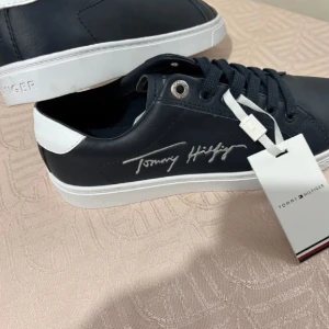 Marinblå sneakers från Tommy Hilfiger - Snygga marinblå sneakers från Tommy Hilfiger med vit logga på sidan och vit detalj vid hälen. Skorna har rund tå, platt vit sula och klassisk snörning. Tillverkade i skinnliknande material och passar perfekt till en clean streetstyle.