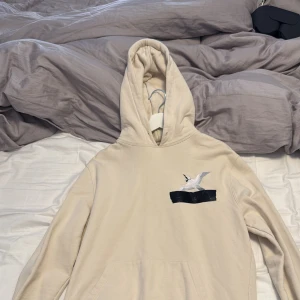 Beige hoodie från Axel Arigato - Snygg beige hoodie från Axel Arigato med broderad fågel och svart detalj på bröstet. Klassisk känguruficka framtill, huva och mjukt material som känns riktigt skönt. Perfekt för dig som gillar stilrena plagg med en twist. Använt nån enstaka gång annars helt ny! 