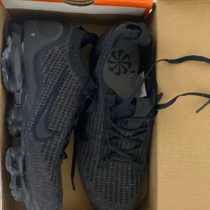 Nike Vapormax Flyknit Svart 42.5 - Säljer ett par svarta Nike Vapormax Flyknit med stickad ovandel och genomskinlig, bubblig sula. Skorna har snörning och diskret Nike-logga på sidan. Perfekta för dig som gillar sportig och modern stil.