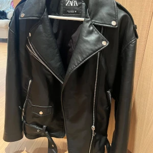 Svart skinnjacka från Zara - Snygg svart skinnjacka från Zara med klassisk bikerstil. Jackan har silvriga dragkedjor, knappar och bälte i midjan. Flera fickor med dragkedja och coola detaljer som ger en edgy vibe. Perfekt för dig som vill ha en tidlös och trendig look.