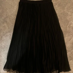 Svart plisserad långkjol - Snygg svart plisserad långkjol med flowig känsla och elastisk midja. Perfekt för dig som gillar en stilren och enkel look med lite extra volym. Kjolen har ett skirt tyg som ger en lätt och luftig vibe.