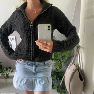 Mörkgrå stickad kofta från Vero Moda - Mysig mörkgrå stickad kofta från Vero Moda med grovt kabelstickat mönster. Koftan har lång ärm, hög krage och en snygg dragkedja med rund detalj. Perfekt för lager-på-lager och chill höst- och vinterstil.