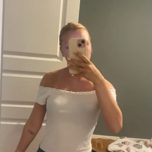 Vit offshoulder topp från Gina Tricot - Supersöt vit offshoulder topp från Gina Tricot med ribbad struktur och små volangdetaljer längs kanten. Sitter tight och har korta ärmar som visar axlarna. Perfekt till jeans eller kjol för en fräsch look.