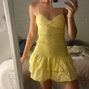 Gul rutig kortklänning med volang - Supersöt gul och vit rutig kortklänning med smockad överdel, rosett framtill och volangig kjol. Klänningen är ärmlös med tunna axelband och har en lätt och luftig känsla, perfekt för varma dagar.